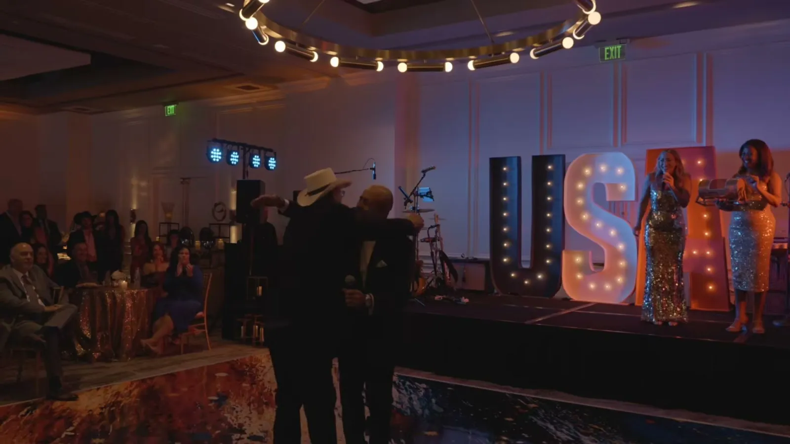 Wide shot of the ballroom at PGA National Resort during the gala, with guests at their tables and two singers on stage beside lit USA marquee letters