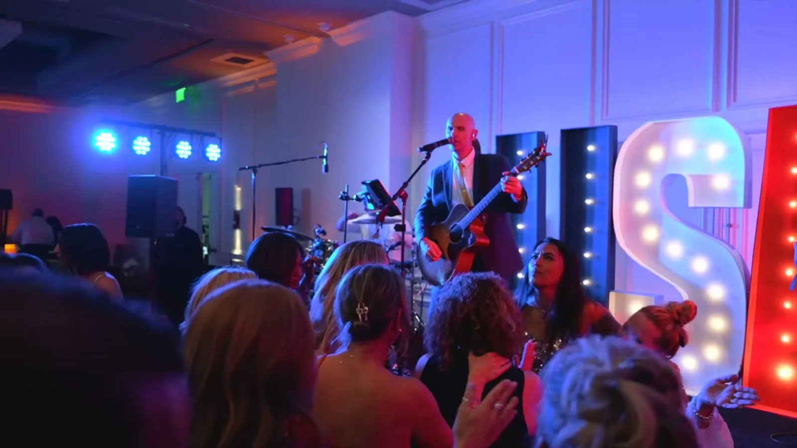 Guests on the dance floor facing the stage as the guitarist sings beside the lit USA marquee letters