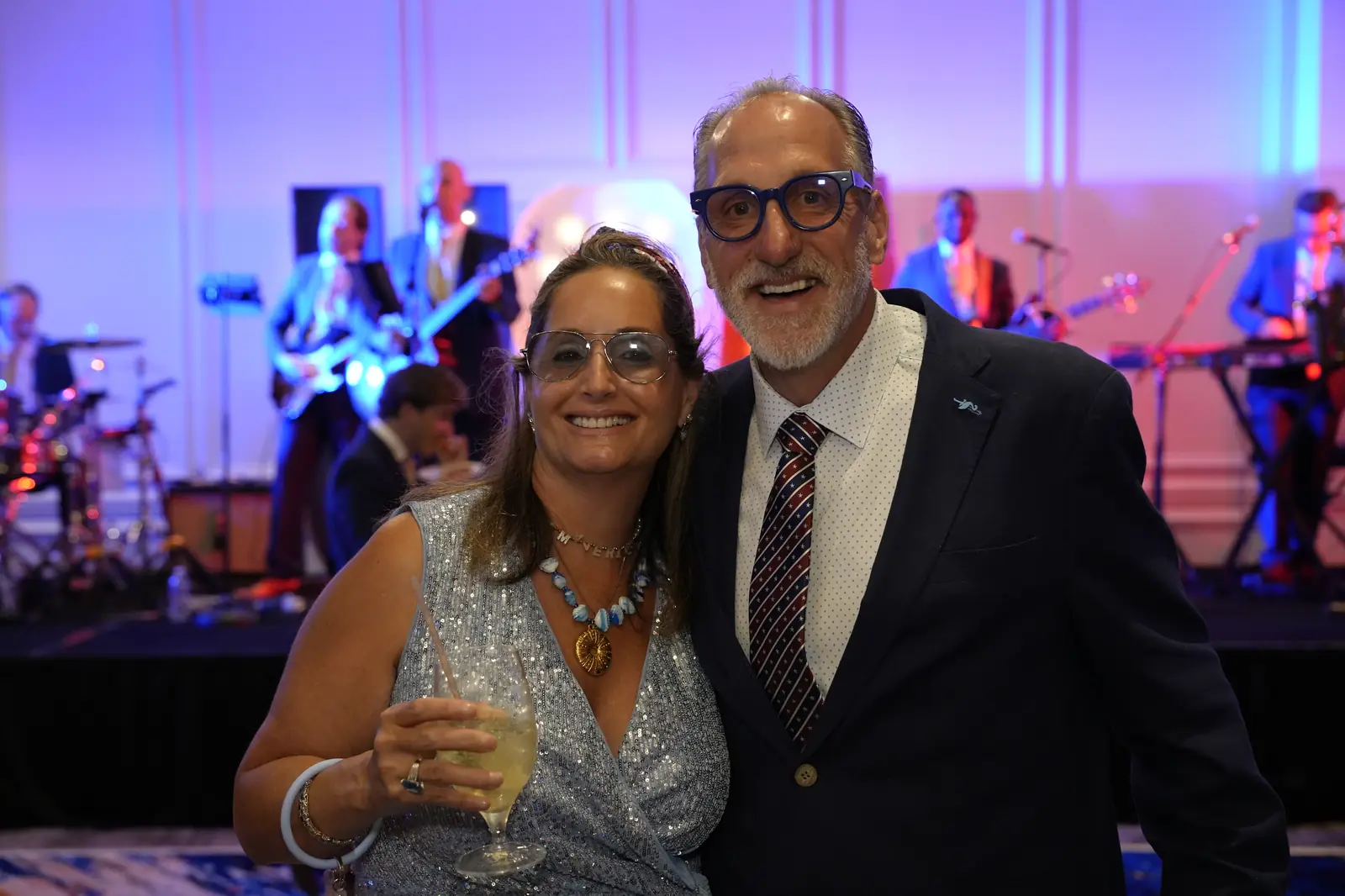 A smiling couple, she raising a drink and he in blue glasses and a striped tie, in front of the band playing on stage