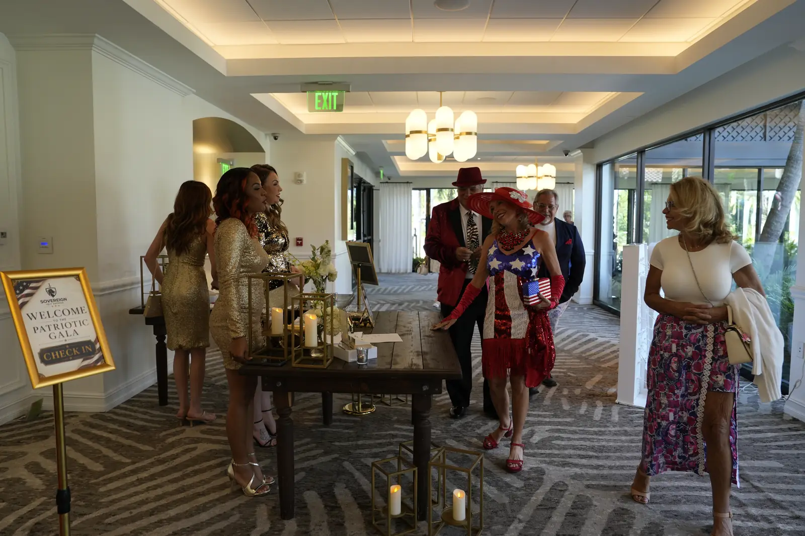 Guests in sequins, a stars-and-stripes dress and a red sun hat arriving at the gala check-in table beside a Welcome to the Patriotic Gala sign at PGA National Resort
