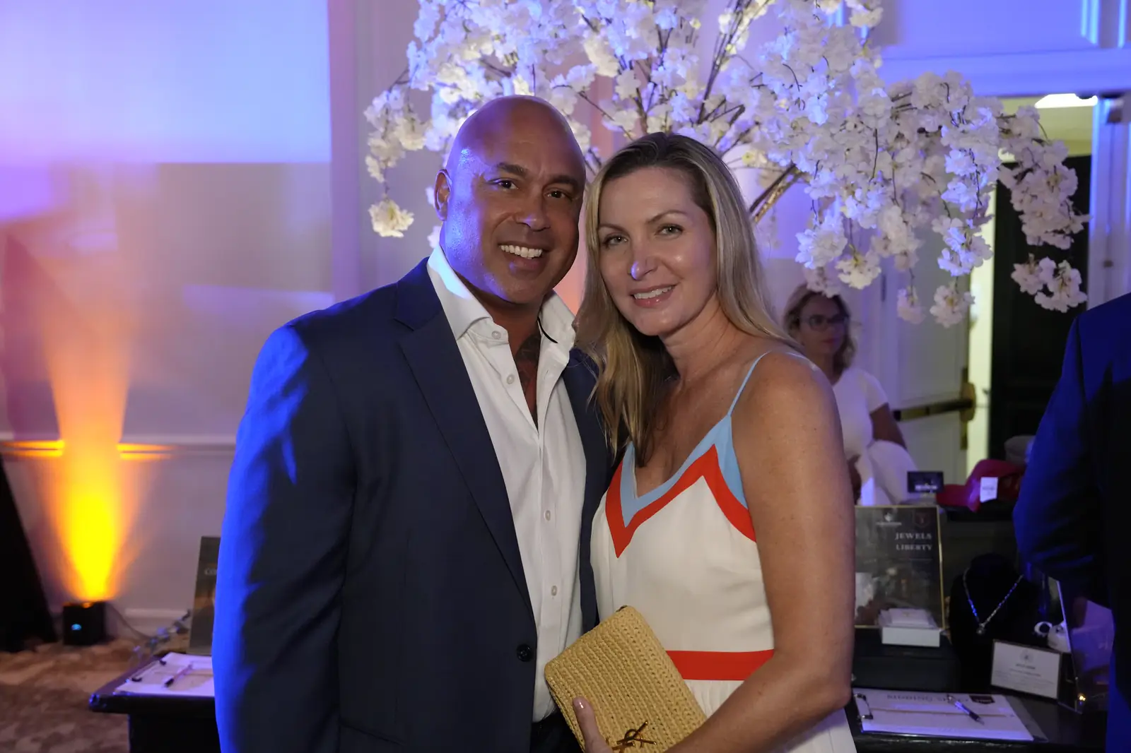 A couple posing in front of a white blossom tree under blue and amber uplighting, beside a display table