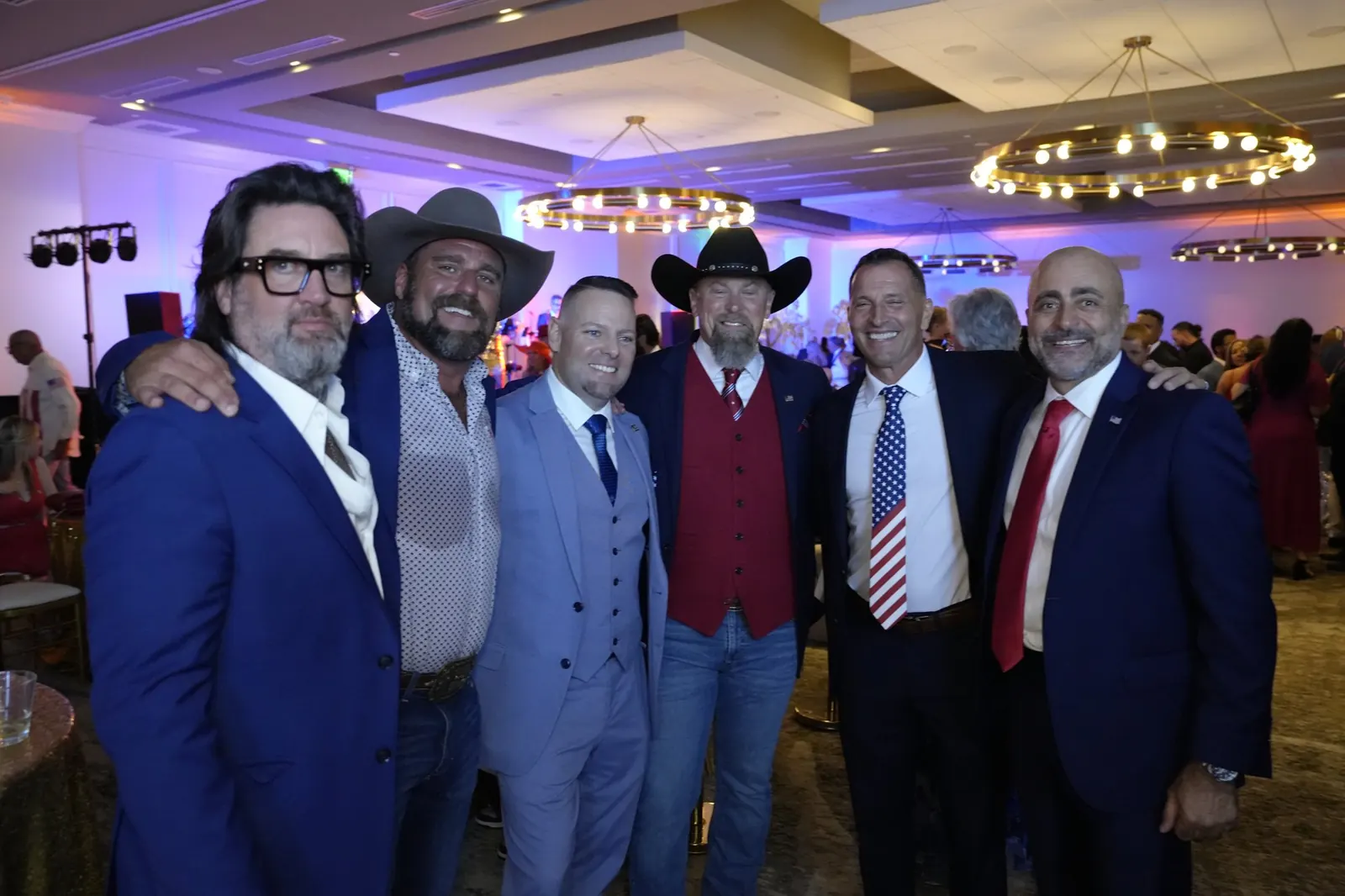 Six men in suits, two of them in cowboy hats, posing arm in arm in the ballroom, with chandeliers and guests behind them