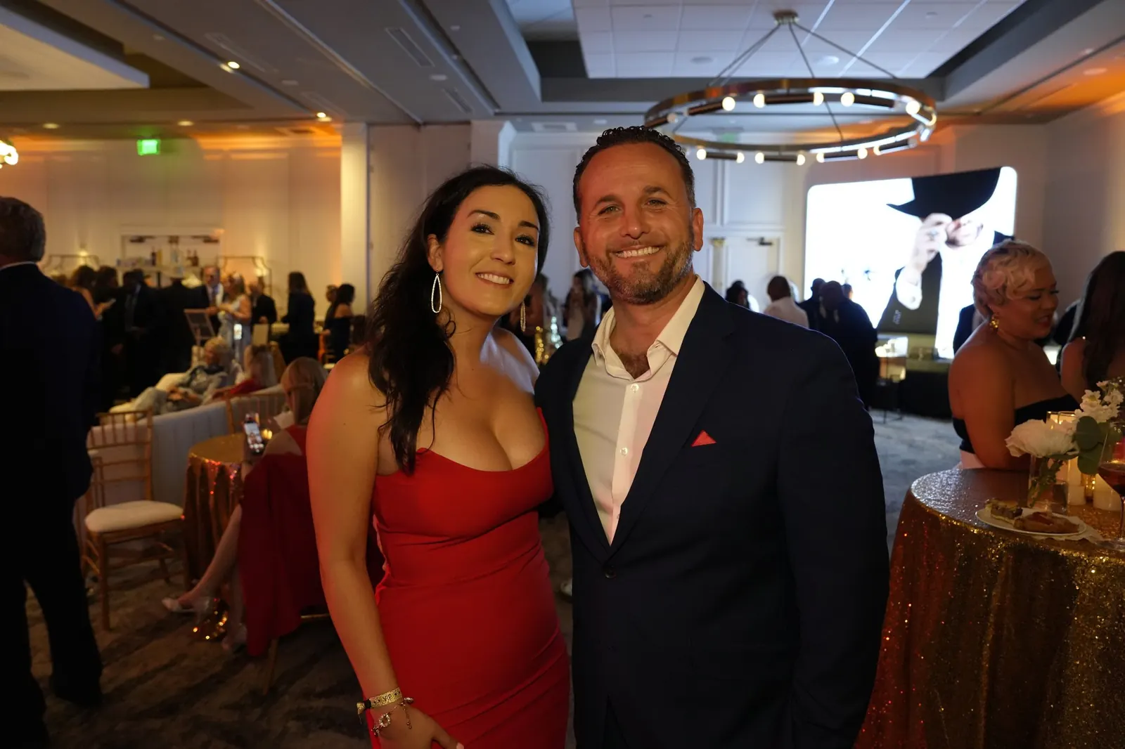 A guest in a red dress and a guest in a navy suit posing among gold-draped tables in the ballroom, with the crowd behind them