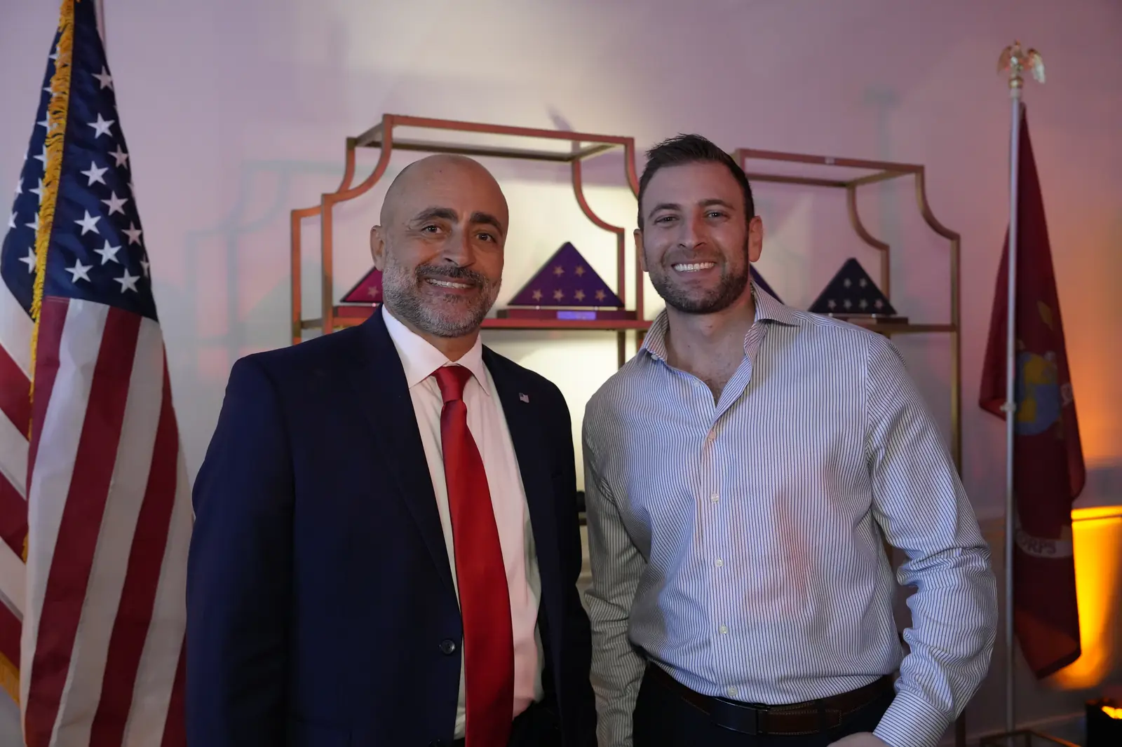 Two smiling men, one in a navy suit and red tie, standing in front of a display of folded American flags in cases beside an American flag