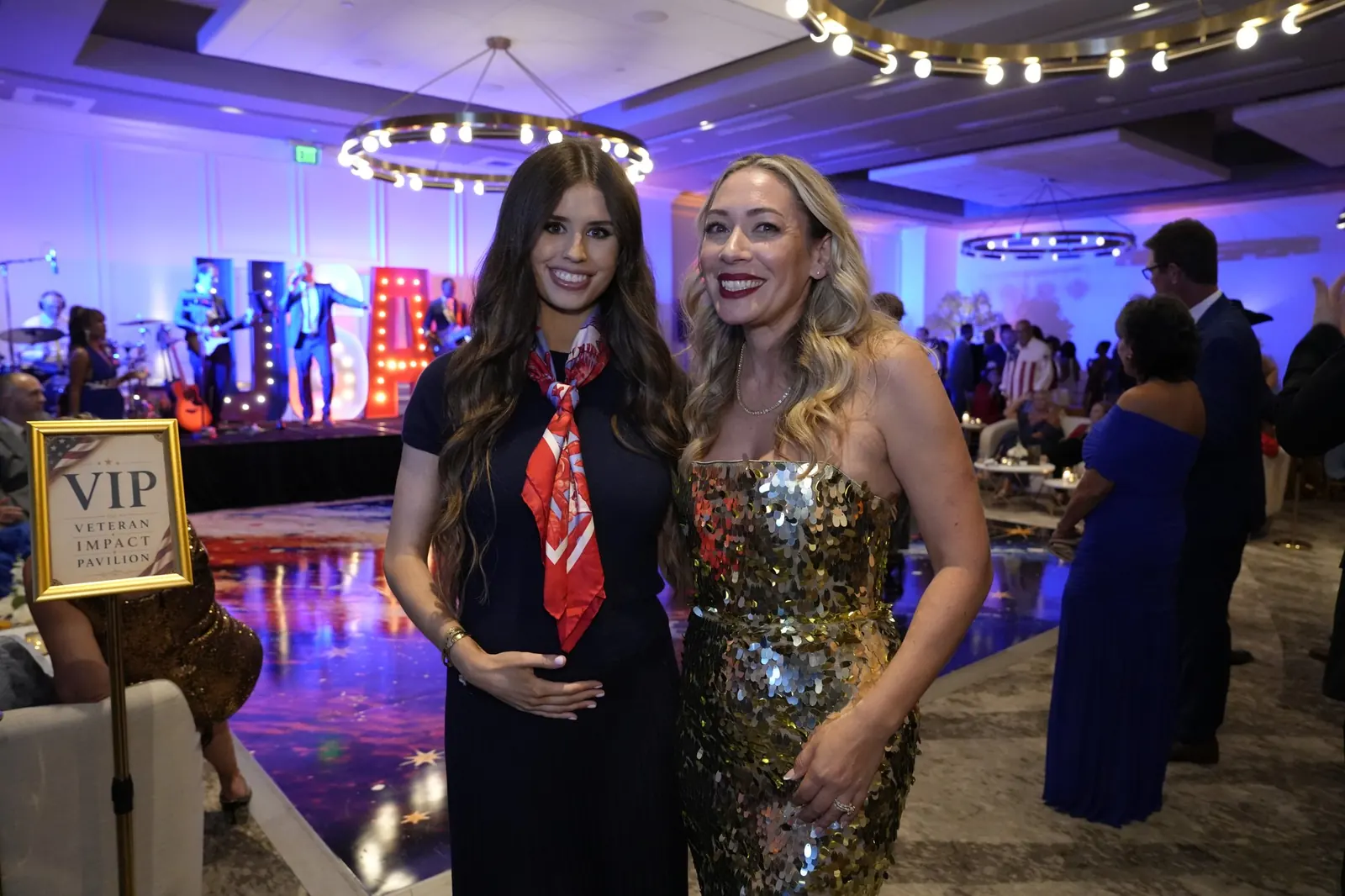 Two gala guests, one in a red scarf and one in a gold sequin dress, posing near the stage and the lit USA marquee letters beside a VIP sign