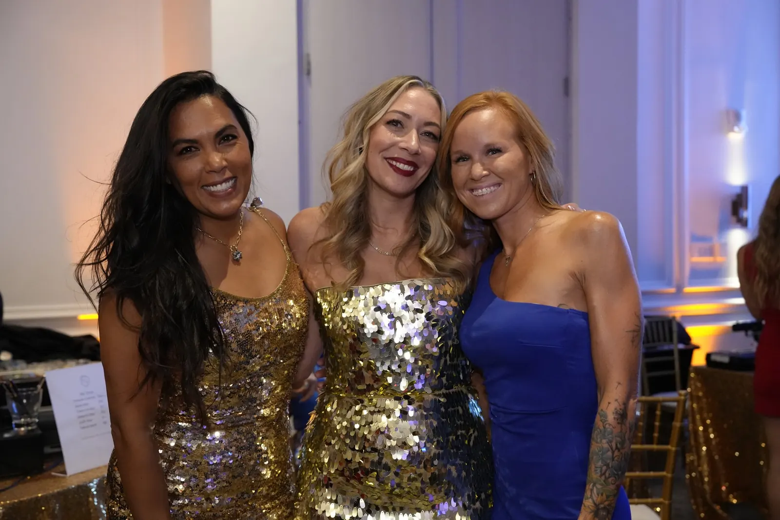 Three gala guests in gold sequin and blue dresses posing together under warm uplighting