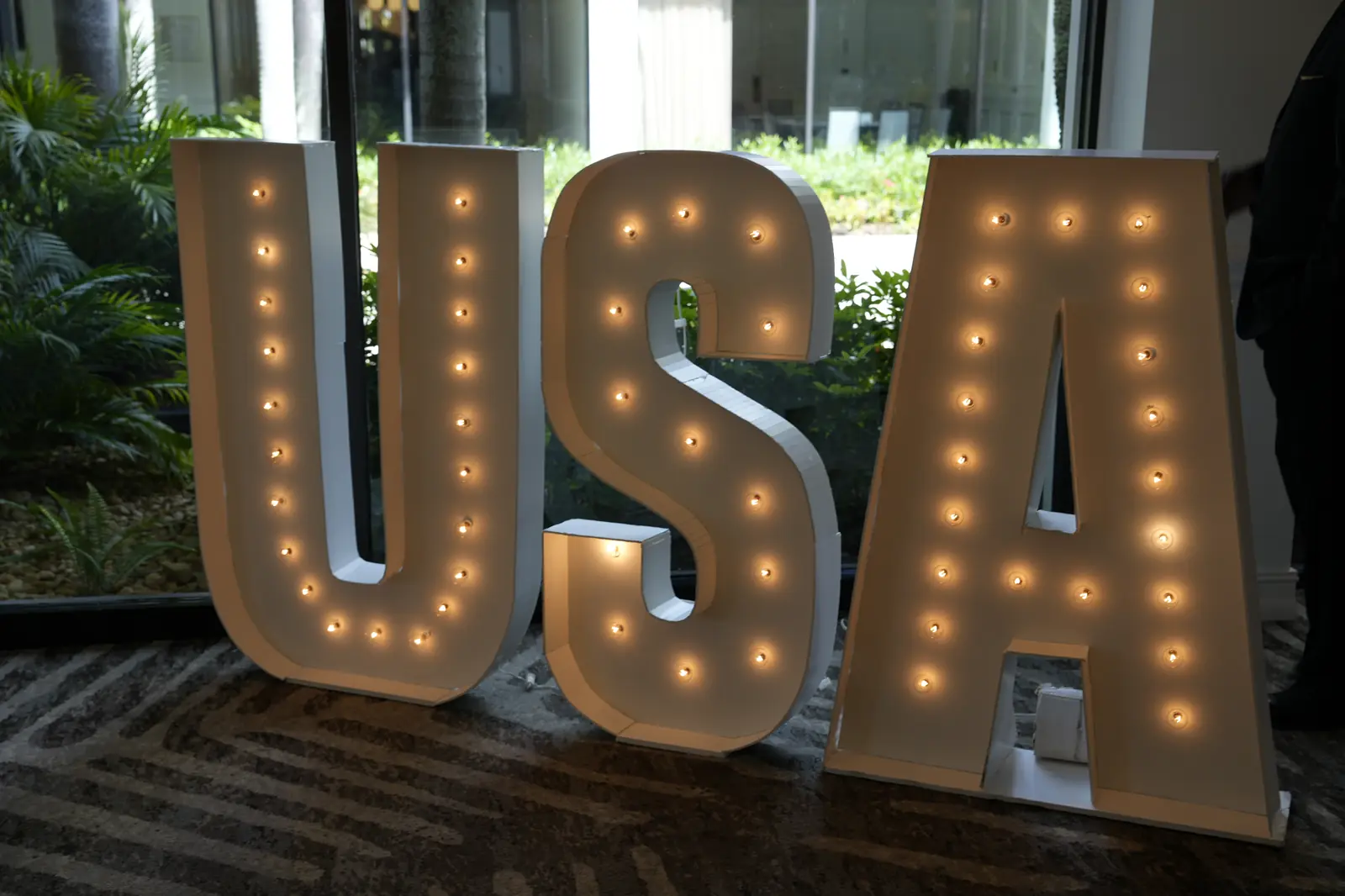 Lit white USA marquee letters standing on the carpet in front of a window onto tropical plants at the gala