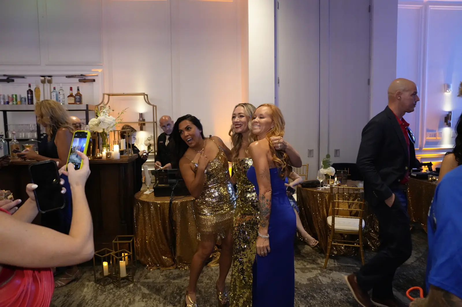 Three guests in gold and blue dresses posing for a friend's phone photo beside the bar while other guests walk past