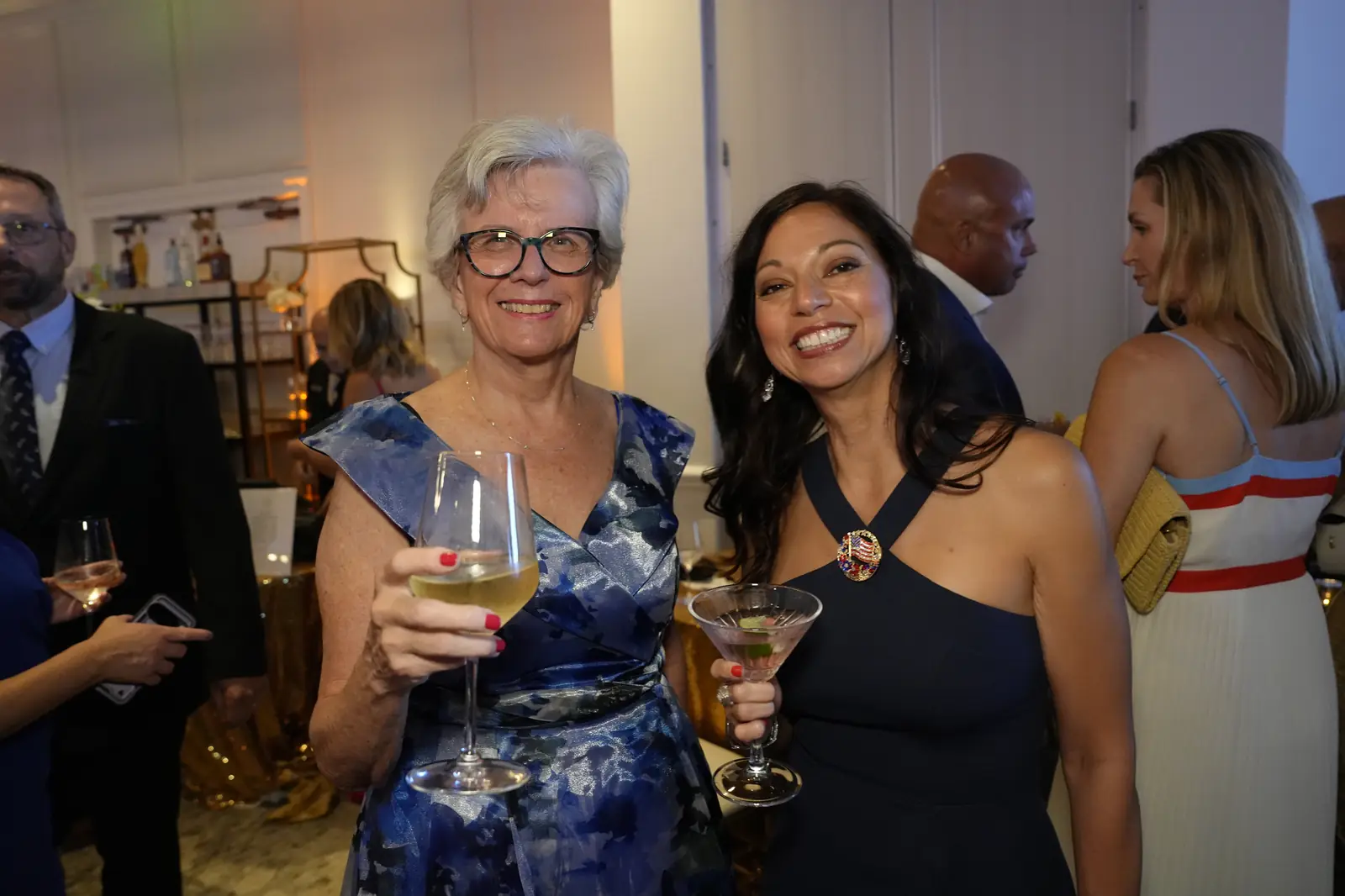Two smiling guests raising a glass of wine and a martini in the busy reception area near the bar