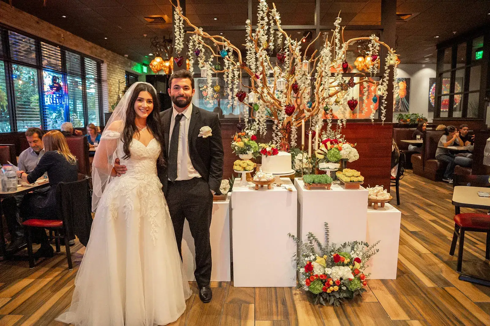 The bride and groom standing by the dessert table, cake and floral centerpiece in a restaurant dining room at their reception