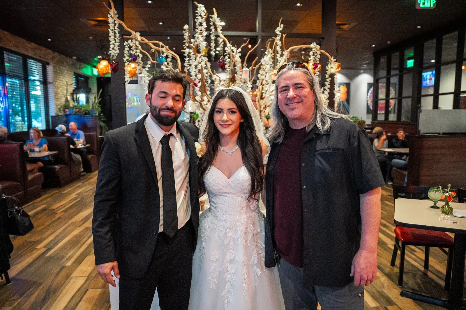 The bride and groom with a guest in front of the blossom tree, the restaurant dining room behind them