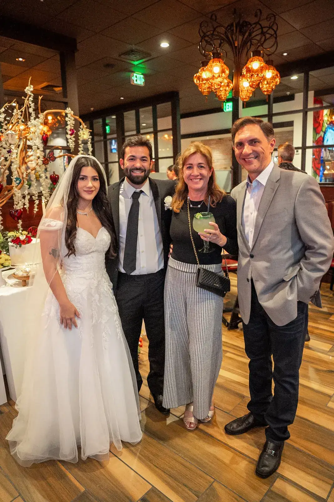 The bride in her veil and the groom standing with two guests, one holding a margarita, beside the dessert table at the reception