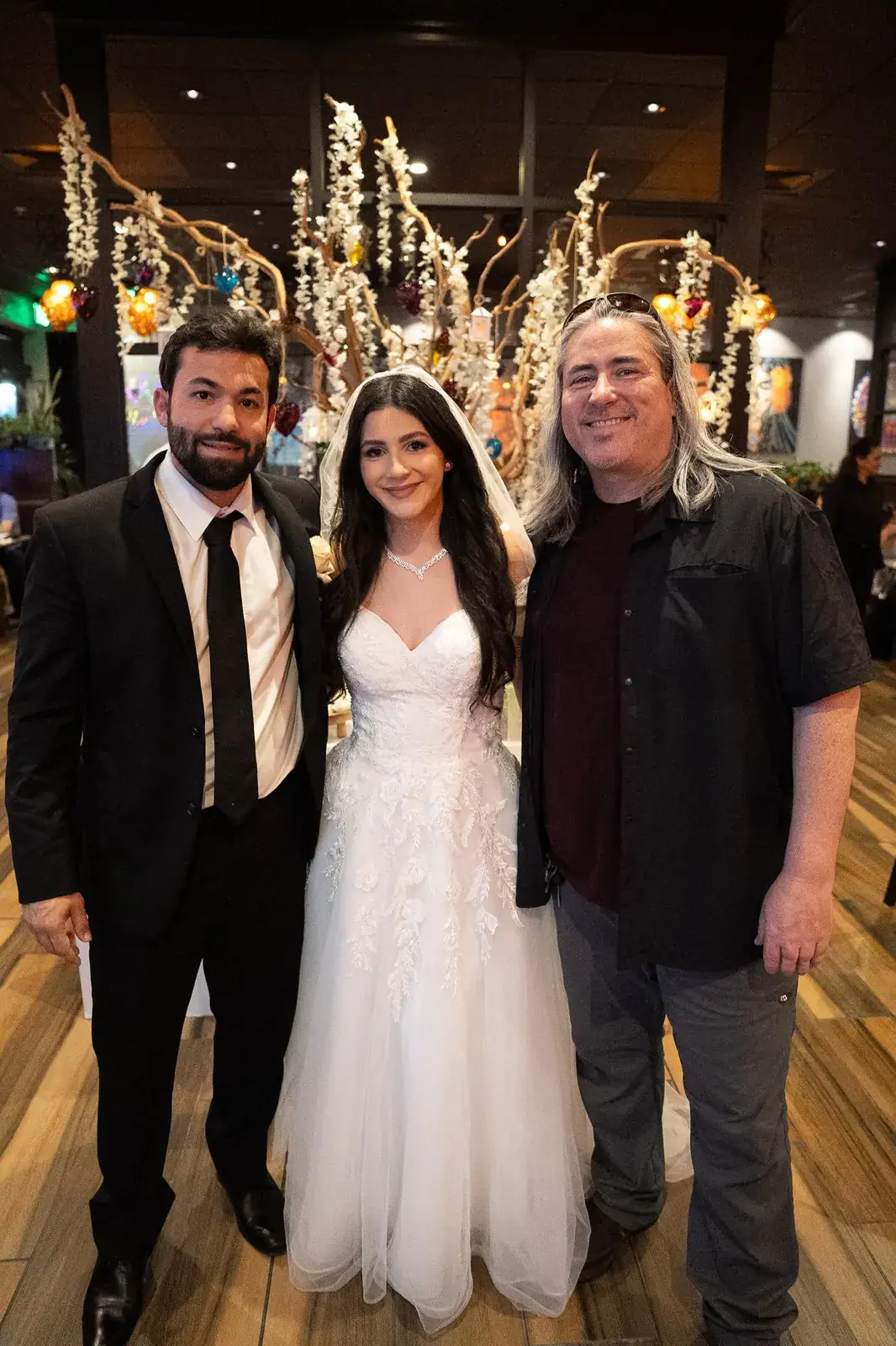 The bride and groom posing with a guest in front of a white blossom tree centerpiece at their wedding reception